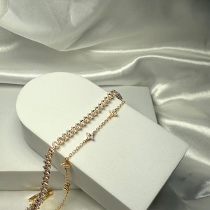 May include: A gold chain bracelet with a delicate design featuring small, clear cubic zirconia stones. The bracelet is layered with a second chain featuring larger, clear cubic zirconia stones.
