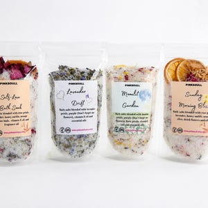 May include: Four clear stand-up pouches containing bath salts with botanical inclusions. Labels read Pinksoull Self-Love Bath Soak, Lavender Drift, Moonlit Garden, and Sunday Morning Bliss, listing ingredients like rose petals, lavender, and dried orange slices.