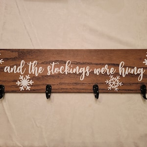 May include: A wooden sign with the text "and the stockings were hung" in white lettering. The sign has four black hooks and white snowflake designs.