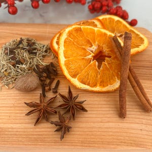 Peut inclure: Une collection d'épices de Noël séchées comprenant deux tranches d'orange, deux bâtons de cannelle, trois étoiles d'anis, une noix de muscade entière, des aiguilles de romarin et des clous de girofle disposés sur une surface en bois clair.