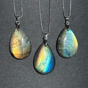 May include: Three labradorite pendants, each with a teardrop shape, hanging on silver chains. The stones show iridescent colours of blue, yellow, and green. The necklaces are simple and elegant, perfect for everyday wear.