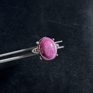 May include: A silver ring with a large, oval, polished pink gemstone. The gemstone is set in a silver band with decorative details. The ring is held by silver tweezers against a dark background.