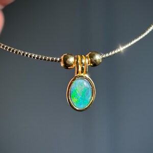May include: A gold necklace with an oval opal pendant. The pendant displays green and blue hues. The necklace has a fine chain and is held up by a hand. The opal pendant is a beautiful piece of jewellery.