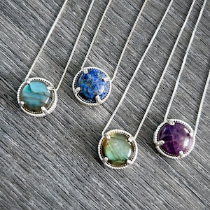 以下が含まれることがあります： 異なる宝石のペンダントが付いた銀のネックレスが4本。ペンダントは丸型で、銀のベゼルセッティングが施されています。宝石はラブラドライト、ラピスラズリ、ラブラドライト、アメジストです。