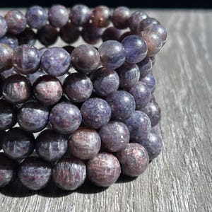 May include: A stack of round, beaded bracelets. The beads are a mix of purple and reddish-brown hues, with a polished, slightly speckled appearance. The bracelets are arranged on a wooden surface, showcasing their color and texture.