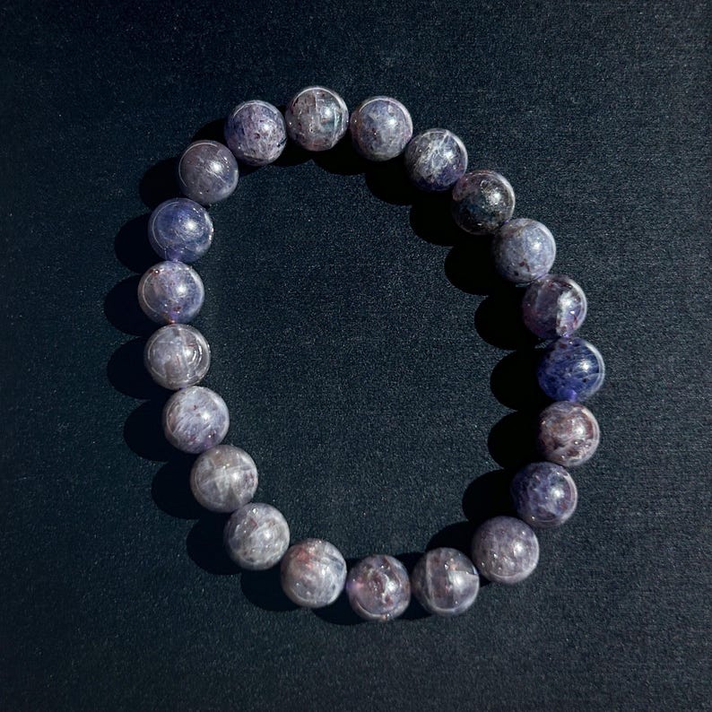 May include: A beaded bracelet composed of round, polished, purple-gray gemstones. The bracelet is displayed on a dark background, with the beads varying in color from light gray to deep purple. The beads are approximately 1 cm in diameter.