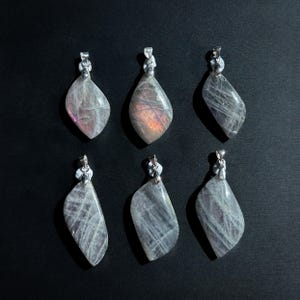 Pu&ograve; includere: Sei ciondoli a forma di goccia con bails color argento. I ciondoli sono grigi traslucidi con striature interne e lampi di rosa e arancione. I ciondoli sono disposti su uno sfondo nero.