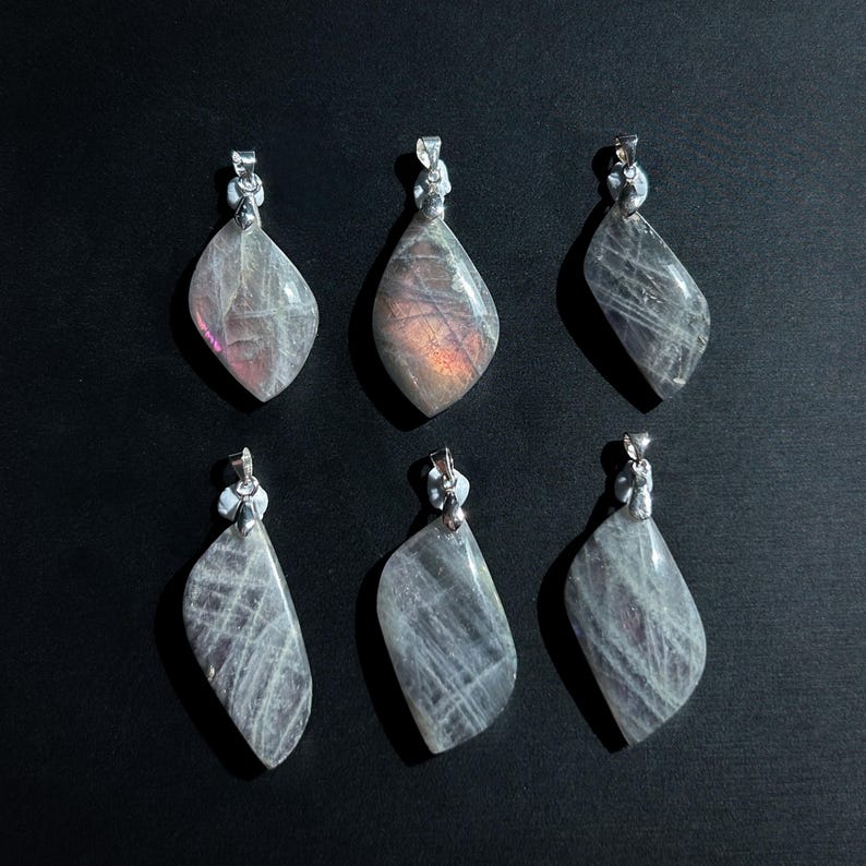Pu&ograve; includere: Sei ciondoli a forma di goccia con bails color argento. I ciondoli sono grigi traslucidi con striature interne e lampi di rosa e arancione. I ciondoli sono disposti su uno sfondo nero.