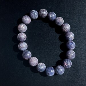 May include: A bracelet made of round, polished beads in shades of purple and gray. The beads have a speckled appearance and are strung together on an elastic band. The bracelet is displayed on a dark background.