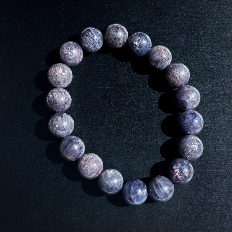 May include: A bracelet made of round, polished beads in shades of purple and gray. The beads have a speckled appearance and are strung together on an elastic band. The bracelet is displayed on a dark background.
