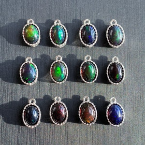 May include: A selection of oval opal pendants with silver frames. The opals exhibit a range of colours, including green, blue, red, and black. Each pendant has a small loop at the top for attaching to a chain or other jewellery component.