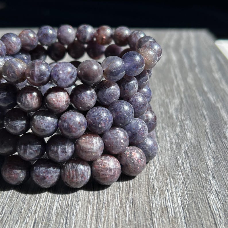 May include: A stack of round, beaded bracelets. The beads are a mix of purple and reddish-brown hues, with a polished, slightly speckled appearance. The bracelets are arranged on a wooden surface, showcasing their color and texture.