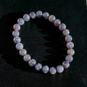 May include: A beaded bracelet featuring round, translucent beads in shades of lavender, purple, and hints of red. The bracelet is strung together, showcasing the natural variations in the stone's color and texture. The bracelet is a piece of jewelry.