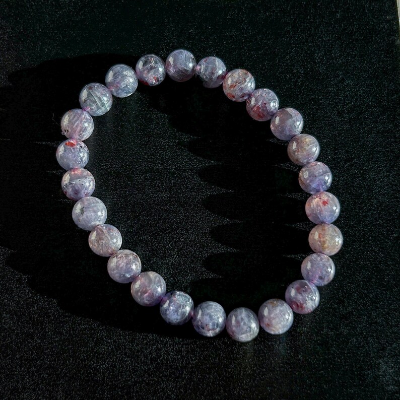 May include: A beaded bracelet featuring round, translucent beads in shades of lavender, purple, and hints of red. The bracelet is strung together, showcasing the natural variations in the stone's color and texture. The bracelet is a piece of jewelry.
