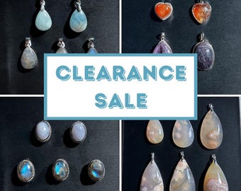 Può includere: Una collezione di pendenti e orecchini con pietre preziose in varie forme e colori, tra cui design a goccia, ovali e a forma di cuore. L'immagine include il testo "CLEARANCE SALE" in un riquadro blu e bianco, su sfondo scuro.