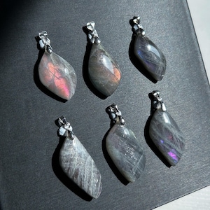 Pu&ograve; includere: Sei ciondoli a forma di goccia con sfumature iridescenti, grigie e viola. Ogni ciondolo ha un'ansa color argento. Le pietre preziose mostrano lampi rosa e arancioni. I ciondoli sono disposti su una superficie scura.