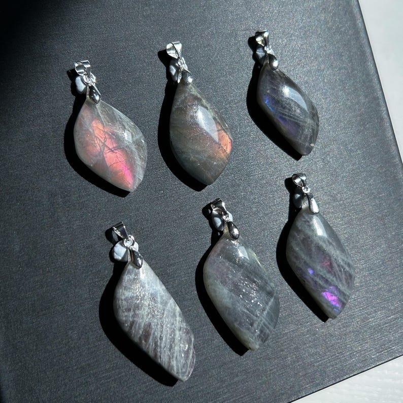Pu&ograve; includere: Sei ciondoli a forma di goccia con sfumature iridescenti, grigie e viola. Ogni ciondolo ha un'ansa color argento. Le pietre preziose mostrano lampi rosa e arancioni. I ciondoli sono disposti su una superficie scura.