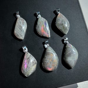 Pu&ograve; includere: Sei ciondoli in labradorite iridescente con bails color argento. I ciondoli hanno una forma a goccia o a foglia, con un mix di colori grigi, viola e blu. I ciondoli sono esposti su una superficie scura.