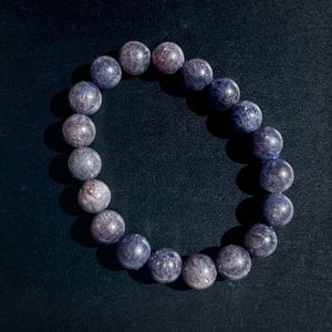 May include: A bracelet featuring round, polished beads in shades of blue and purple. The beads have a speckled appearance, with hints of red and white. The bracelet is strung on a clear elastic band, and is displayed against a dark background.