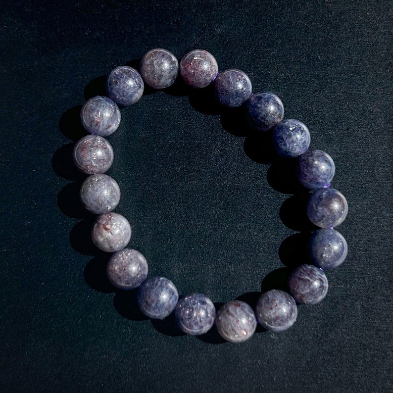 May include: A bracelet featuring round, polished beads in shades of blue and purple. The beads have a speckled appearance, with hints of red and white. The bracelet is strung on a clear elastic band, and is displayed against a dark background.