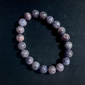 May include: A beaded bracelet featuring round, polished beads in shades of purple and reddish-brown. The bracelet is made of natural gemstones and is designed to be worn around the wrist. The beads have a marbled appearance.