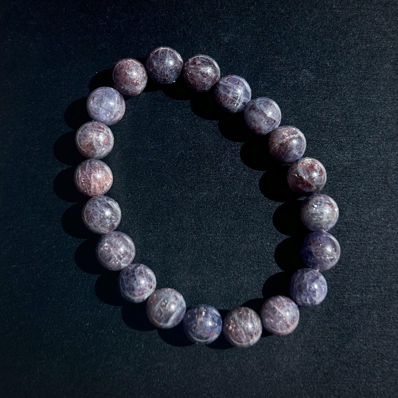 May include: A beaded bracelet featuring round, polished beads in shades of purple and reddish-brown. The bracelet is made of natural gemstones and is designed to be worn around the wrist. The beads have a marbled appearance.