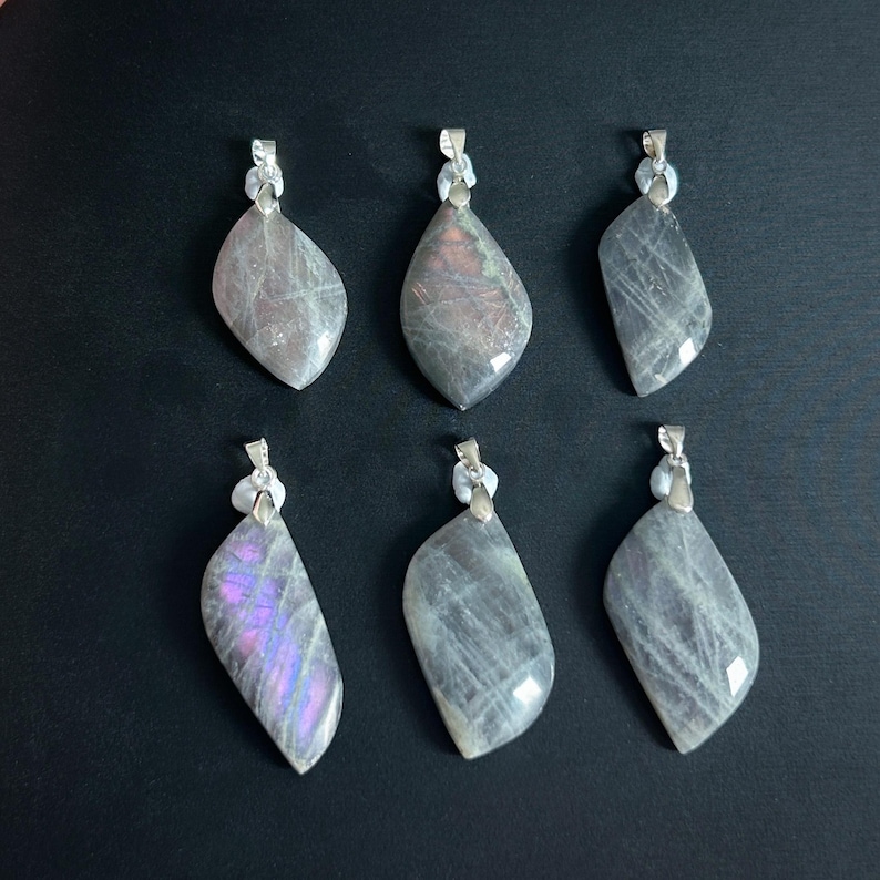 Pu&ograve; includere: Sei ciondoli con pietre preziose con bails color argento sono esposti su uno sfondo scuro. I ciondoli sono a forma di goccia, con un mix di colori grigi, viola e iridescenti. Ogni ciondolo ha un motivo e una consistenza unici.