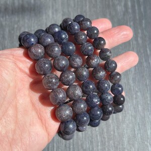 May include: A strand of dark blue-grey gemstone beads. The beads are round and have a polished, reflective surface. The beads are held in a hand, showcasing their color and texture. The beads are strung together, forming a bracelet or necklace.