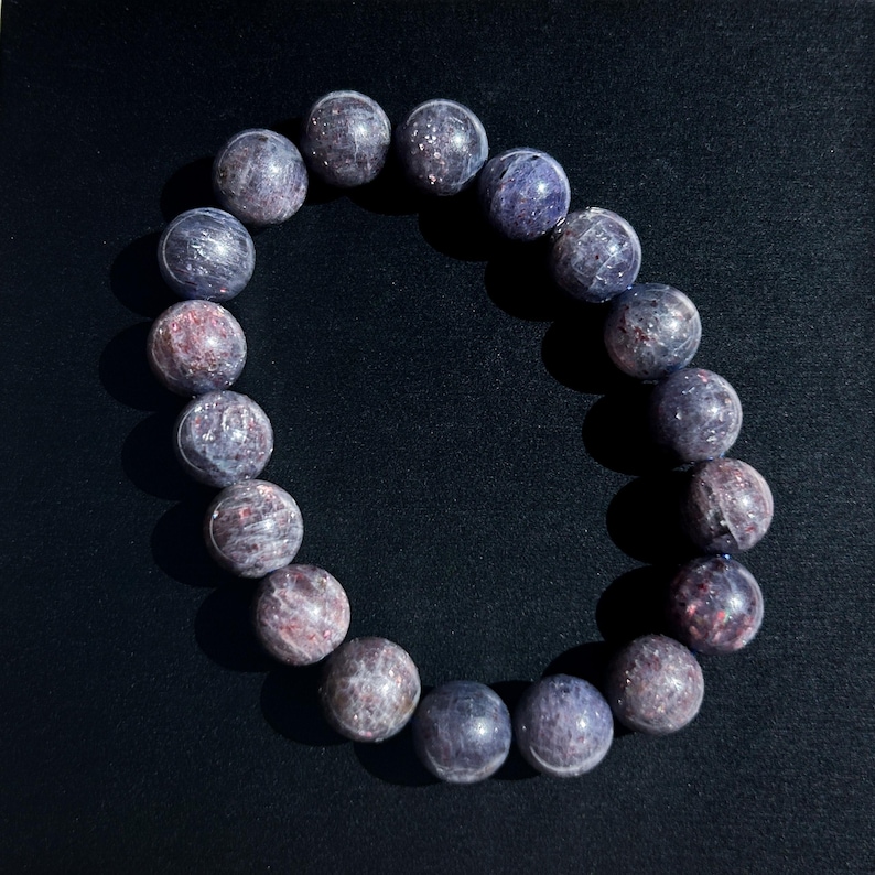 May include: A beaded bracelet featuring round, polished stones in shades of purple, gray, and hints of red. The bracelet is made of multiple spherical beads strung together, creating a circular shape. The beads have a speckled appearance and a glossy finish.