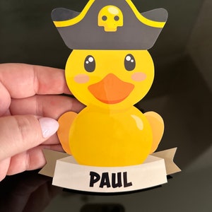May include: A yellow rubber duck sticker featuring a dark gray pirate hat with a yellow skull icon. The duck has an orange beak and black eyes. Below the duck is a light beige banner with the name PAUL printed in bold black capital letters.