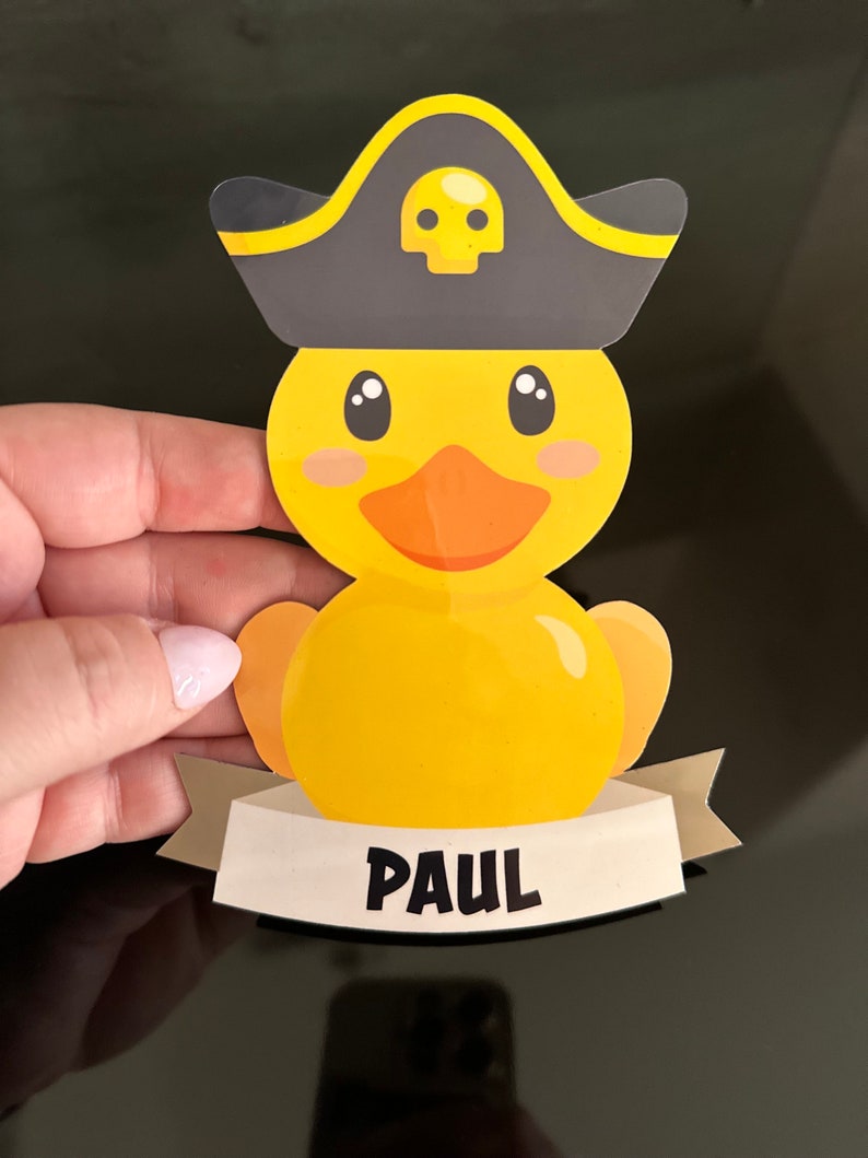 May include: A yellow rubber duck sticker featuring a dark gray pirate hat with a yellow skull icon. The duck has an orange beak and black eyes. Below the duck is a light beige banner with the name PAUL printed in bold black capital letters.