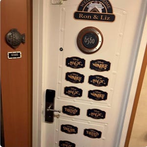 May include: A white door with a porthole-style window displaying the number 6560. The door is decorated with Disney cruise line themed stickers, including "Magic", "Wonder", "Fantasy", and "Dream". A welcome sign reads "Ron & Liz".