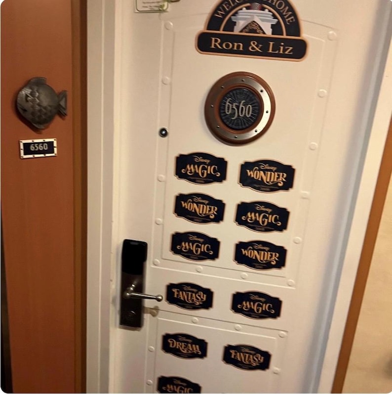 May include: A white door with a porthole-style window displaying the number 6560. The door is decorated with Disney cruise line themed stickers, including "Magic", "Wonder", "Fantasy", and "Dream". A welcome sign reads "Ron & Liz".