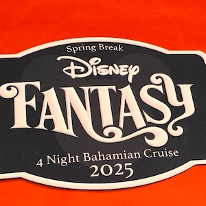 May include: A black and white Disney Fantasy cruise sign with the text "Spring Break", "Disney", "FANTASY", "4 Night Bahamian Cruise", and "2025". The sign has a white border and is set against an orange background.