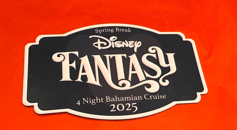 May include: A black and white Disney Fantasy cruise sign with the text "Spring Break", "Disney", "FANTASY", "4 Night Bahamian Cruise", and "2025". The sign has a white border and is set against an orange background.