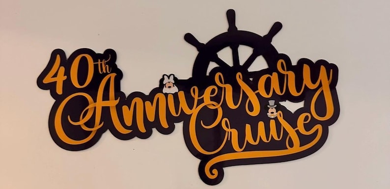 Op de afbeelding: Een zwart en oranje bord met de tekst 40th Anniversary Cruise in een gestileerd scriptlettertype. Het ontwerp bevat een zwart icoon van een scheepsroer en twee kleine stripfiguurhoofdjes die tussen de woorden zijn geplaatst.
