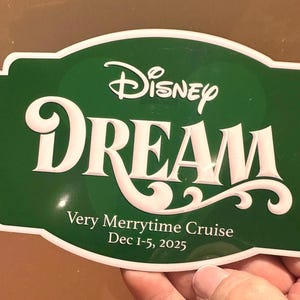 May include: A green sign with white trim and text that reads "Disney DREAM" in a stylized font. Below, it says "Very Merrytime Cruise Dec 1-5, 2025". The sign is held by a hand.