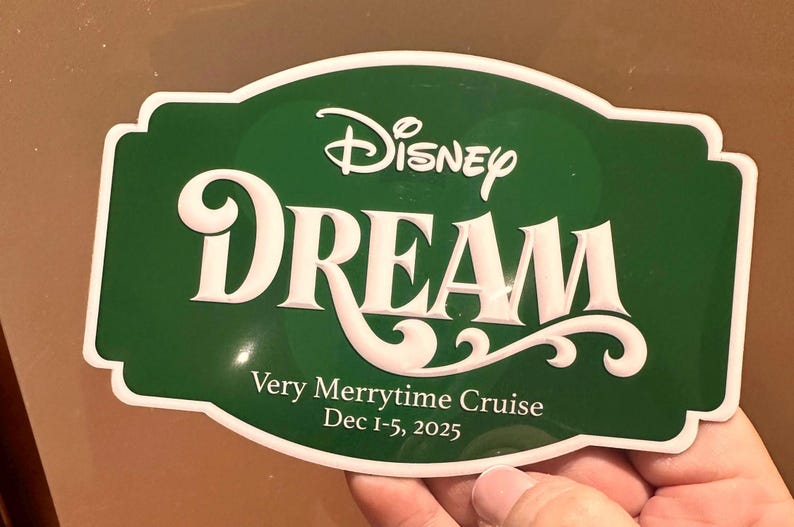 May include: A green sign with white trim and text that reads "Disney DREAM" in a stylized font. Below, it says "Very Merrytime Cruise Dec 1-5, 2025". The sign is held by a hand.
