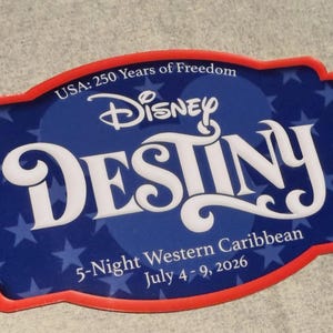 May include: A rectangular Disney Destiny souvenir with a red border. The blue center features white text that reads "DESTINY" and "Disney". The top text reads "USA: 250 Years of Freedom". The bottom text reads "5-Night Western Caribbean July 4-9, 2026".