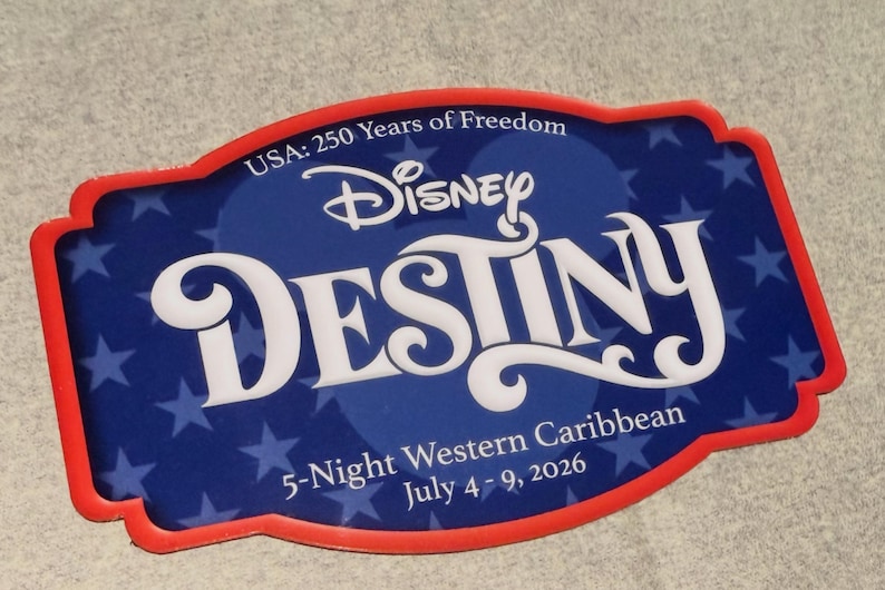 May include: A rectangular Disney Destiny souvenir with a red border. The blue center features white text that reads "DESTINY" and "Disney". The top text reads "USA: 250 Years of Freedom". The bottom text reads "5-Night Western Caribbean July 4-9, 2026".