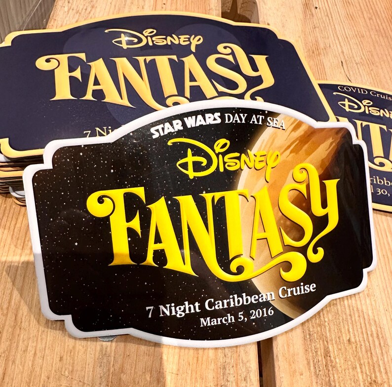 May include: Several Disney Fantasy cruise signs. The signs have a navy blue and black background with yellow text that reads "Disney FANTASY". One sign is for a "7 Night Caribbean Cruise" on March 5, 2016, with "Star Wars Day at Sea".