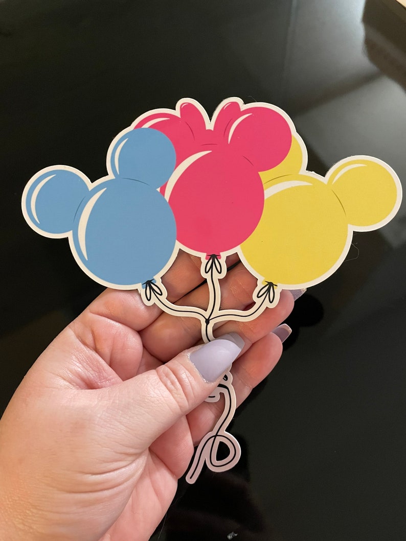 Pode incluir: Tr&ecirc;s bal&otilde;es em forma de Mickey Mouse nas cores azul, rosa e amarelo. Os bal&otilde;es est&atilde;o amarrados com um cord&atilde;o.