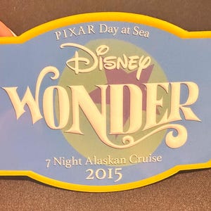 May include: A blue and yellow Disney Cruise Line souvenir with the text "PIXAR Day at Sea" and "Disney WONDER". The souvenir is for a "7 Night Alaskan Cruise 2015". The souvenir is held by a person.