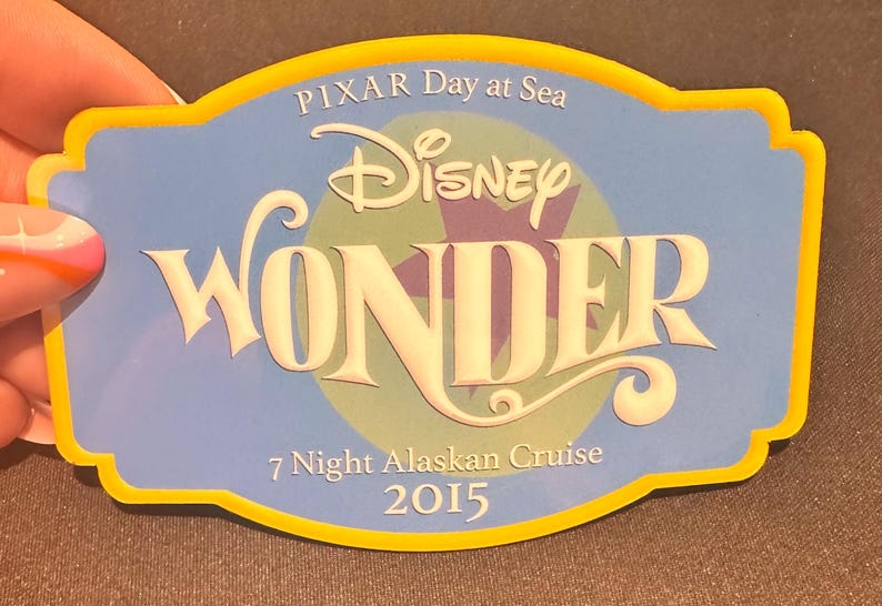 May include: A blue and yellow Disney Cruise Line souvenir with the text "PIXAR Day at Sea" and "Disney WONDER". The souvenir is for a "7 Night Alaskan Cruise 2015". The souvenir is held by a person.