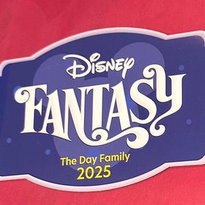 May include: A blue and white Disney Fantasy sign with the text "Disney Fantasy" in a stylized font. Below the main text are the words "The Day Family 2025". The sign is set against a red background.