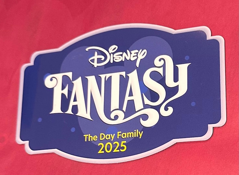 May include: A blue and white Disney Fantasy sign with the text "Disney Fantasy" in a stylized font. Below the main text are the words "The Day Family 2025". The sign is set against a red background.