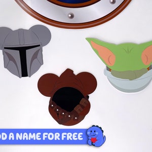 May include: Three character head cutouts with ears, featuring a gray Mandalorian helmet, a green Baby Yoda, and a brown hooded figure. The text "ADD A NAME FOR FREE" is at the bottom of the image.