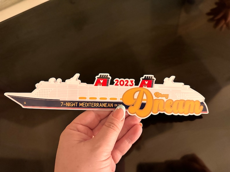 May include: A white and navy blue cruise ship sticker with the text "Disney Dream" in orange script. The sticker also includes the text "2023" and "7-Night Mediterranean". The ship has red and black Mickey Mouse ears.