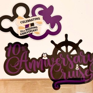 Op de afbeelding: Twee decoratieve borden in zwart en paars. Het bovenste bord toont twee overlappende muisoren-silhouetten met een banner met de tekst CELEBRATING 10TH WEDDING ANNIVERSARY. Het onderste bord toont een scheepsroer en de tekst 10th Anniversary Cruise.