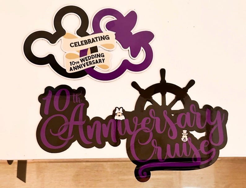 Op de afbeelding: Twee decoratieve borden in zwart en paars. Het bovenste bord toont twee overlappende muisoren-silhouetten met een banner met de tekst CELEBRATING 10TH WEDDING ANNIVERSARY. Het onderste bord toont een scheepsroer en de tekst 10th Anniversary Cruise.
